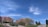 Rows of red-brick apartment buildings are seen against a bright blue sky with scattered white clouds. In the foreground, there are several trees and well-maintained shrubs, along with a grassy area with stone landscaping and a set of stairs leading up a hill.