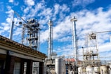 Multiple tall, metallic structures and pipelines are positioned against a backdrop of a blue sky with scattered clouds. The industrial setting suggests a refinery or factory, complete with various tanks and support beams.