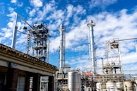 Multiple tall, metallic structures and pipelines are positioned against a backdrop of a blue sky with scattered clouds. The industrial setting suggests a refinery or factory, complete with various tanks and support beams.