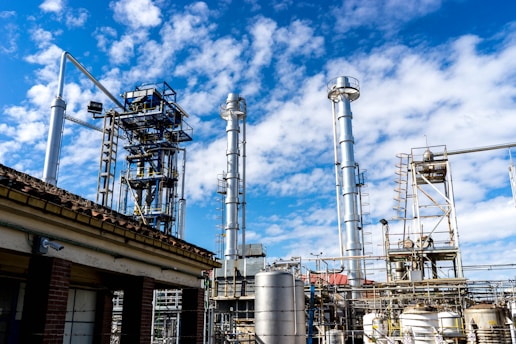 Multiple tall, metallic structures and pipelines are positioned against a backdrop of a blue sky with scattered clouds. The industrial setting suggests a refinery or factory, complete with various tanks and support beams.