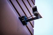 A security camera mounted on a wall with visible cables. The wall has a textured surface with horizontal lines, and the background is a clear blue sky.