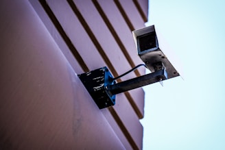 A security camera mounted on a wall with visible cables. The wall has a textured surface with horizontal lines, and the background is a clear blue sky.