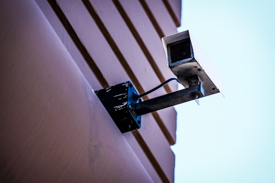 A security camera mounted on a wall with visible cables. The wall has a textured surface with horizontal lines, and the background is a clear blue sky.