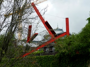 Large red steel beams form an abstract structure amidst a green, leafy environment. A construction crane and a distinctive building with an ornate roof are visible in the background.