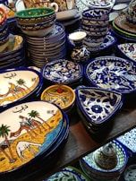 A collection of colorful, intricately designed ceramic dishware featuring artistic patterns and illustrations. The items include plates and bowls with vibrant hues of blue, yellow, and white, and illustrations depicting traditional scenes.