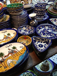 A collection of colorful, intricately designed ceramic dishware featuring artistic patterns and illustrations. The items include plates and bowls with vibrant hues of blue, yellow, and white, and illustrations depicting traditional scenes.