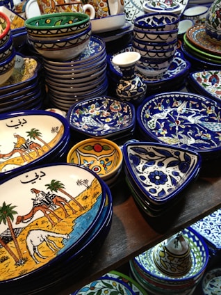 A collection of colorful, intricately designed ceramic dishware featuring artistic patterns and illustrations. The items include plates and bowls with vibrant hues of blue, yellow, and white, and illustrations depicting traditional scenes.