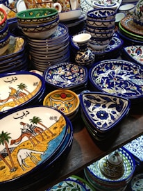 A collection of colorful, intricately designed ceramic dishware featuring artistic patterns and illustrations. The items include plates and bowls with vibrant hues of blue, yellow, and white, and illustrations depicting traditional scenes.