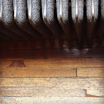 An ornate metal radiator with detailing is positioned above a worn, wooden floor. The radiator has vertically aligned sections with intricate swirl designs. The wooden floorboards show signs of wear and scratches.
