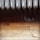 An ornate metal radiator with detailing is positioned above a worn, wooden floor. The radiator has vertically aligned sections with intricate swirl designs. The wooden floorboards show signs of wear and scratches.