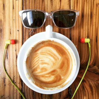 A cup of coffee with latte art is placed on a wooden table. Above the cup, a pair of sunglasses is positioned, and two brightly colored earphones flank the cup, creating a playful arrangement.