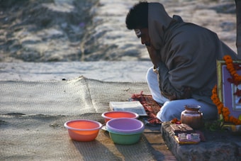 A person wrapped in a shawl is sitting on a mat with several bowls and a metal pot nearby. The person appears to be focused on an open book in front of them. The setting suggests a calm and meditative atmosphere, possibly during a religious or spiritual ritual. A garland of orange flowers is also visible beside the person.