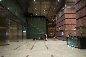 A spacious, modern lobby with tall ceilings and marble floors. The walls have a blend of dark green and reddish-brown marble. A reception desk is to the right with plants in vases atop it. There is seating in the middle and two people standing nearby. Elevators with metallic doors line the left side.