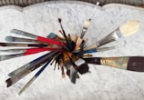 A collection of various paintbrushes is arranged in a circular formation from an overhead perspective. The brushes, featuring different sizes and shapes, display handles of multiple colors, including red, blue, and brown. The background consists of a white marbled surface, providing a neutral contrast to the vivid brush colors.