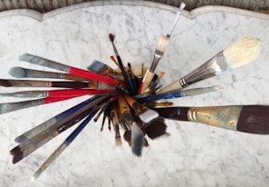 A collection of various paintbrushes is arranged in a circular formation from an overhead perspective. The brushes, featuring different sizes and shapes, display handles of multiple colors, including red, blue, and brown. The background consists of a white marbled surface, providing a neutral contrast to the vivid brush colors.