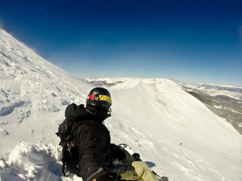A person wearing a helmet and a backpack is sitting on a snowy mountain slope, possibly preparing to ski or snowboard. The landscape features a vast expanse of snow, with rugged mountain peaks in the distance under a clear blue sky.