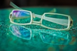 A pair of rectangular eyeglasses with metal frames lies on a textured glass surface, creating a reflection. The lenses have a subtle purple tint, and the background features a soft blur of green and blue hues.