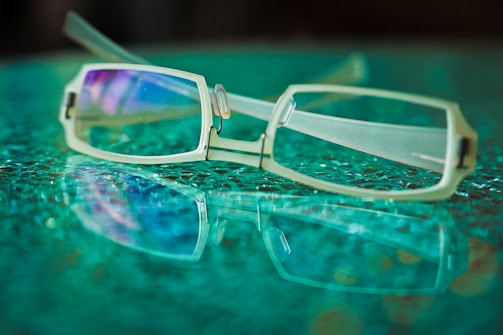 A pair of rectangular eyeglasses with metal frames lies on a textured glass surface, creating a reflection. The lenses have a subtle purple tint, and the background features a soft blur of green and blue hues.