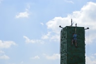 A person is climbing a tall, green artificial rock climbing wall with a safety harness under a clear blue sky with scattered fluffy clouds.