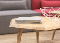 A contemporary wooden coffee table with a geometric design on the top surface. Two identical books titled 'The Unsplash Book' are stacked on the table. A white, wireframe decorative bowl sits next to the books. In the background, part of a gray sofa with a red cushion is partially visible.