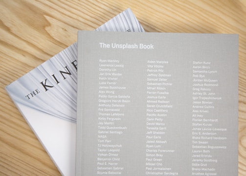 Two books are placed on a light wooden surface. The top book is titled 'The Unsplash Book' and features a list of names printed in white on a light gray cover. Part of the title 'THE KIN' is visible on the book underneath, with a partially visible white cover and black text.