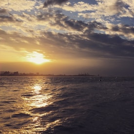The image captures a serene view of the ocean with a golden sunset on the horizon. Light reflects beautifully off the water, creating a peaceful and calm atmosphere. The sky is partially cloudy, adding texture and depth to the scene. In the background, a faint silhouette of a distant city skyline can be observed.