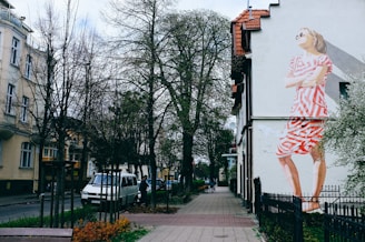 A city street scene features buildings flanking both sides of a narrow sidewalk with bare trees lining the path. On the right side, a large mural of a woman in a geometric red and white dress is painted on the side of a building. A white van is parked on the street, and a few people are visible in the background.