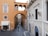 A narrow cobblestone street lined with historic buildings featuring arched windows and doors. The architecture includes an overhead pedestrian bridge connecting two buildings, with a soft orange wall and an intricate window design. A few people are walking along the street, and a shop with an awning reads 'La Veneta' near the entrance.