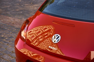 A close-up of the rear section of a red Volkswagen car with a clear reflection of a building on its shiny surface. The Volkswagen logo and the word 'Kafer' are visible on the back of the vehicle. The car is parked on a cobblestone street.