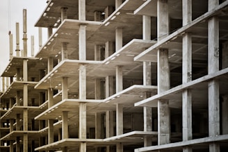 Concrete structures of an unfinished multi-story building with visible floors and supporting pillars. The image captures the skeletal framework, showing repetitive geometric patterns and an industrial atmosphere.