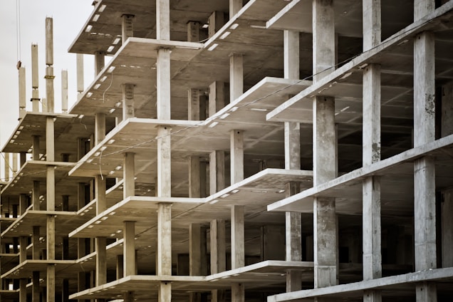 Concrete structures of an unfinished multi-story building with visible floors and supporting pillars. The image captures the skeletal framework, showing repetitive geometric patterns and an industrial atmosphere.