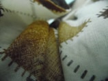 Fabrics displayed with a close-up view of a textured surface, featuring patterns with brown and black stitching against a white background. The material appears to be layered, creating a sense of depth.