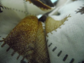 Fabrics displayed with a close-up view of a textured surface, featuring patterns with brown and black stitching against a white background. The material appears to be layered, creating a sense of depth.