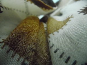 Fabrics displayed with a close-up view of a textured surface, featuring patterns with brown and black stitching against a white background. The material appears to be layered, creating a sense of depth.