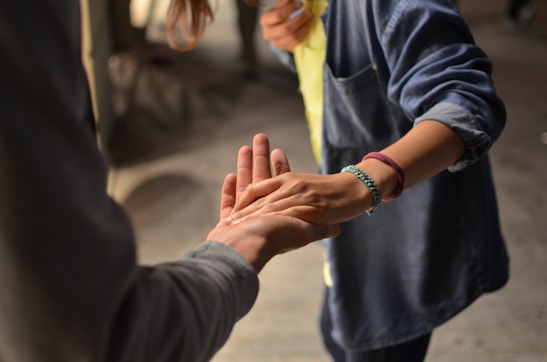 Two people are gently holding hands, suggesting a moment of connection or support. One person is wearing a denim shirt and bracelets, and the setting appears to be casual and intimate.