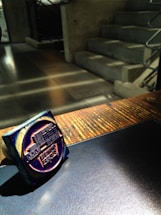 A dark blue ink stamp is resting on a wooden surface with sunlight casting shadows. The background features a concrete staircase with metallic railings, adding an industrial feel to the environment.