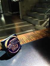 A dark blue ink stamp is resting on a wooden surface with sunlight casting shadows. The background features a concrete staircase with metallic railings, adding an industrial feel to the environment.