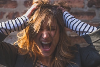 A person pulling their hair with both hands, mouth open wide, possibly in a scream or shout. The individual is wearing a striped long-sleeve shirt under a dark jacket. The background consists of a brick wall.