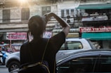 A person with a backpack is taking a photo of a street scene. The surroundings include a mix of vehicles, a motorcycle, and a car under the warm glow of sunlight. The backdrop consists of older buildings with visible signs in a foreign language, creating an urban atmosphere.