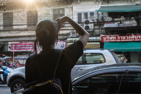 A person with a backpack is taking a photo of a street scene. The surroundings include a mix of vehicles, a motorcycle, and a car under the warm glow of sunlight. The backdrop consists of older buildings with visible signs in a foreign language, creating an urban atmosphere.