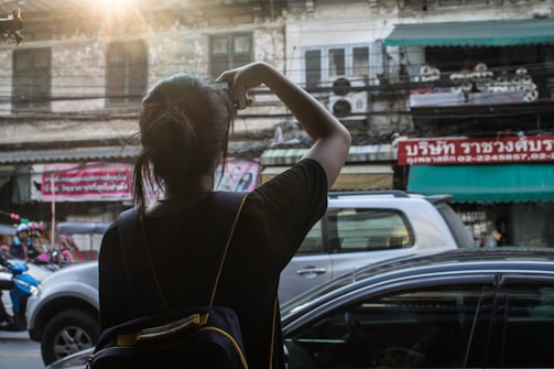 A person with a backpack is taking a photo of a street scene. The surroundings include a mix of vehicles, a motorcycle, and a car under the warm glow of sunlight. The backdrop consists of older buildings with visible signs in a foreign language, creating an urban atmosphere.