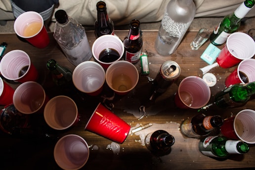 An assortment of red plastic cups, various beer bottles, a vodka bottle, a lighter, a few scattered cigarette butts, and spilled liquid are spread across a wooden table. The scene suggests a party or casual gathering with drinks and smoking items.