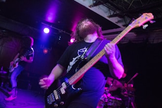 A musician passionately plays an electric bass guitar on stage. The stage is dimly lit with colorful lighting, including purple and blue hues. Two other band members are visible in the background, playing their instruments.