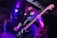 A musician passionately plays an electric bass guitar on stage. The stage is dimly lit with colorful lighting, including purple and blue hues. Two other band members are visible in the background, playing their instruments.