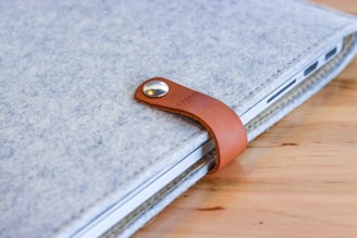 A close-up view of a device, possibly a laptop, enclosed in a gray felt sleeve with a brown leather strap fastened with a metal button. The item rests on a wooden surface.