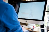 A person wearing a blue shirt is working on a desktop computer with a large screen displaying code. The environment appears to be an office space with natural light coming in from a window. The scene includes a keyboard, mouse, and some desk accessories like pens and notebooks.