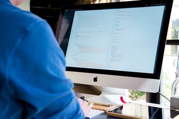 A person wearing a blue shirt is working on a desktop computer with a large screen displaying code. The environment appears to be an office space with natural light coming in from a window. The scene includes a keyboard, mouse, and some desk accessories like pens and notebooks.