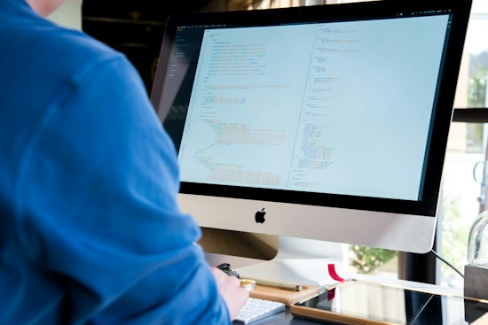A person wearing a blue shirt is working on a desktop computer with a large screen displaying code. The environment appears to be an office space with natural light coming in from a window. The scene includes a keyboard, mouse, and some desk accessories like pens and notebooks.