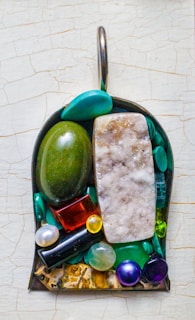 A pendant featuring a variety of gemstones in different shapes and colors, including a large green oval stone, a rectangular crystal with a frosted texture, and several round pearls. The stones are arranged in a metal frame with a hook at the top.