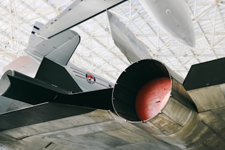 The image depicts a close-up view of a jet aircraft's engine and exhaust area. The aircraft is metallic and appears to be housed in a large hangar or museum with a grid-like ceiling structure. Part of an insignia or logo is visible on the aircraft's body.
